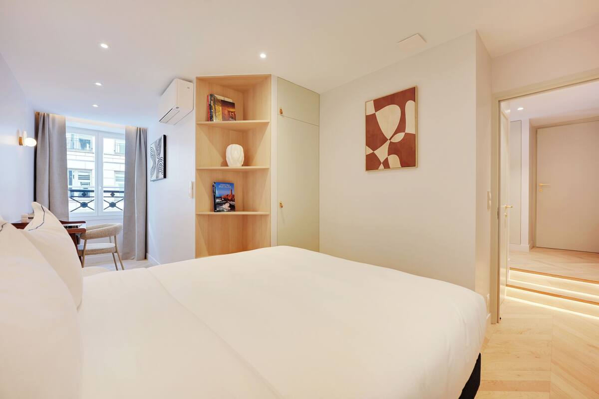 A modern hotel room with a large bed, a wooden bookshelf, a desk, and a view of the city through the window.