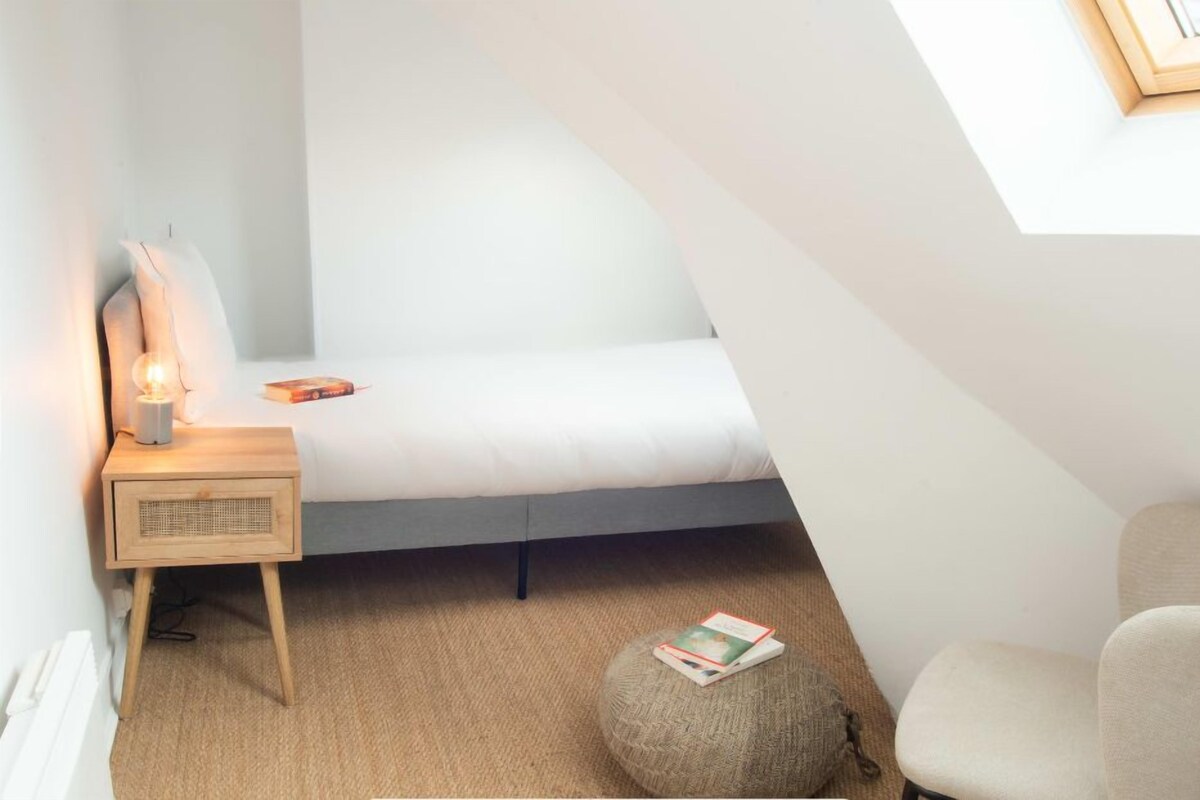 A cozy loft bedroom with a sloped ceiling, a small bedside table with a lamp, a comfortable chair, and a cushioned ottoman.