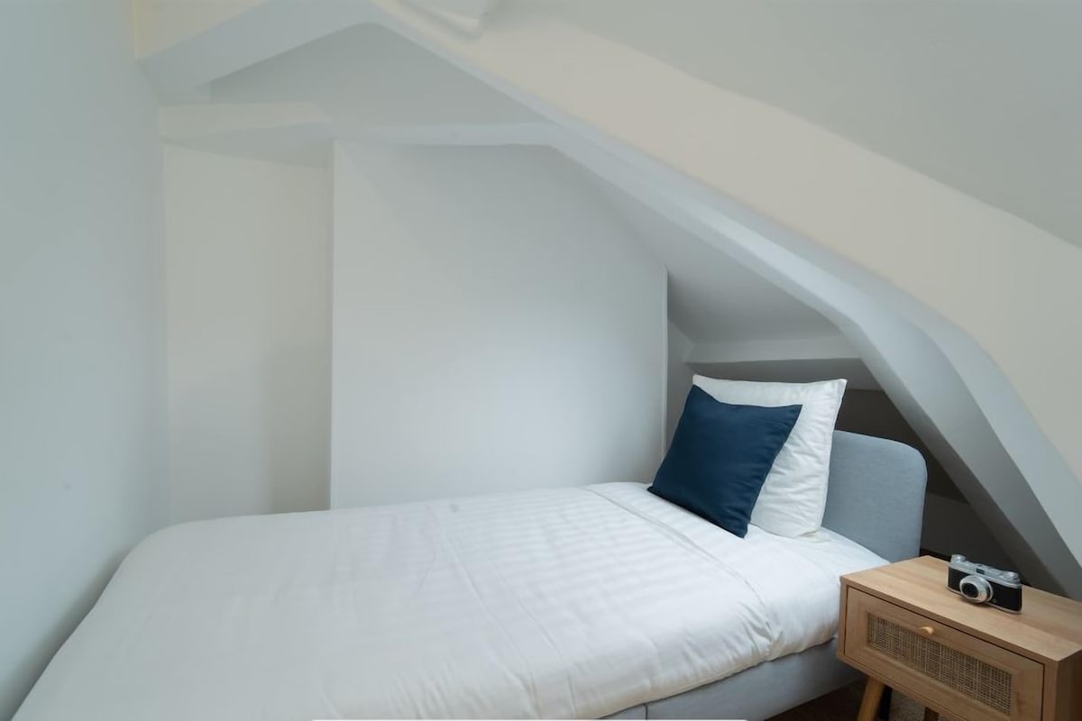 A small, single-bed room with a sloped ceiling, a wooden nightstand, and a camera on it.