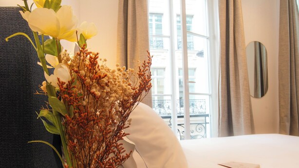 A hotel room with a bed, a vase with flowers, a bedside table, a window with curtains, and a view of buildings outside. - Aparthotel Quartier Libre Madeleine (Paris)