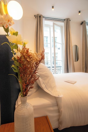 A hotel room with a bed, a vase with flowers, a bedside table, a window with curtains, and a view of buildings outside. - Aparthotel Quartier Libre Madeleine (Paris)