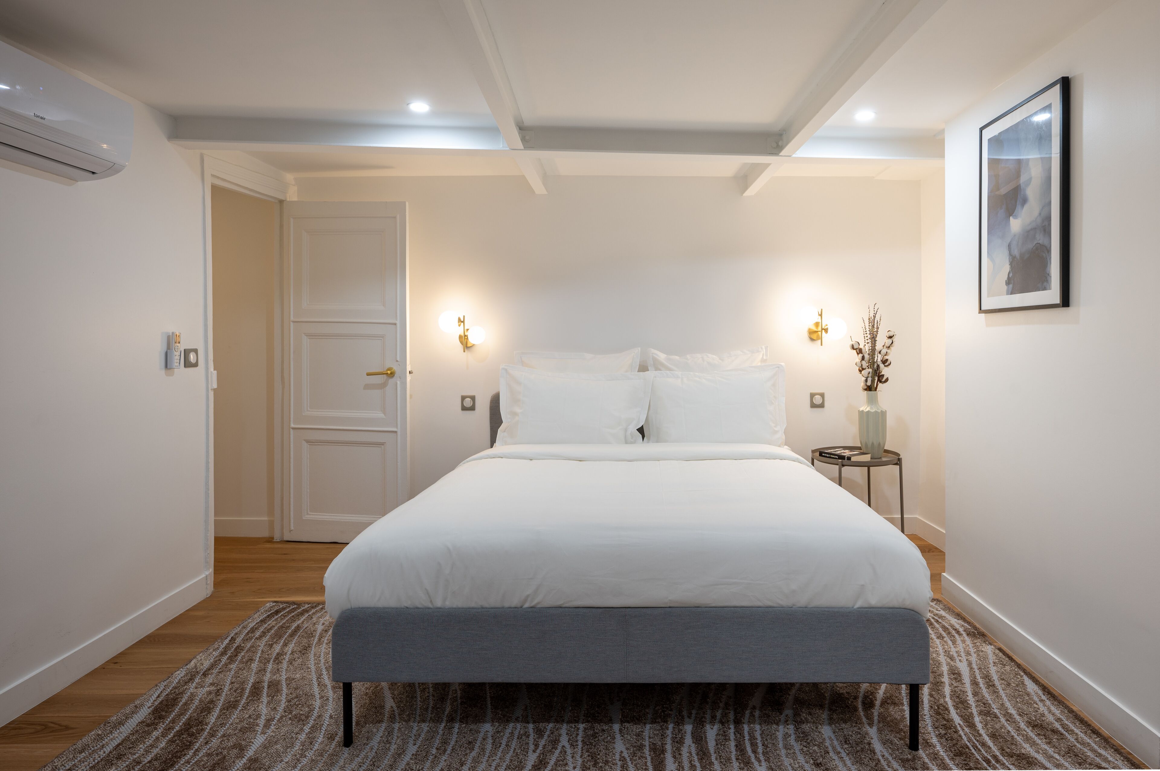A modern bedroom with a large bed, a bedside table with a vase, and a door leading to another room.