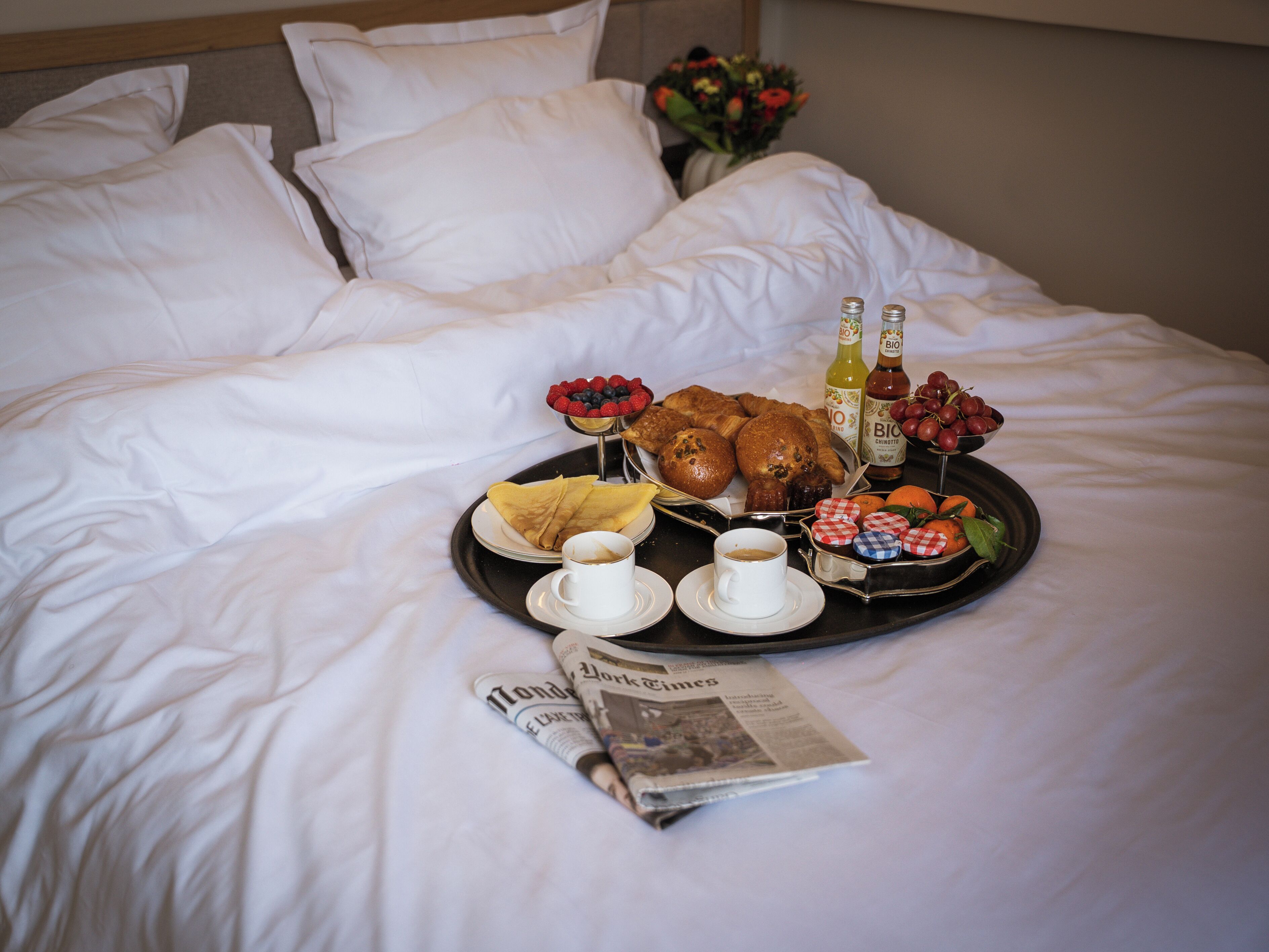 A neatly made bed with white linens, a tray of breakfast items including pastries, fruits, and beverages, and a newspaper on the bed.