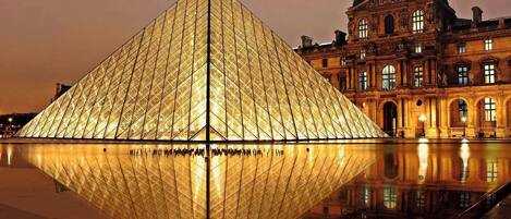 Una gran estructura piramidal iluminada con su reflejo en el agua, junto a un edificio histórico de arquitectura clásica.