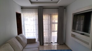 Una sala de estar con un sofá color beige, un tapete azul, un televisor en la pared y una ventana con cortinas traslúcidas.