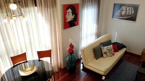 Una sala de estar con una mesa redonda de comedor, dos sillas, un sofá con cojines, un jarrón con flores y dos cuadros en las paredes.