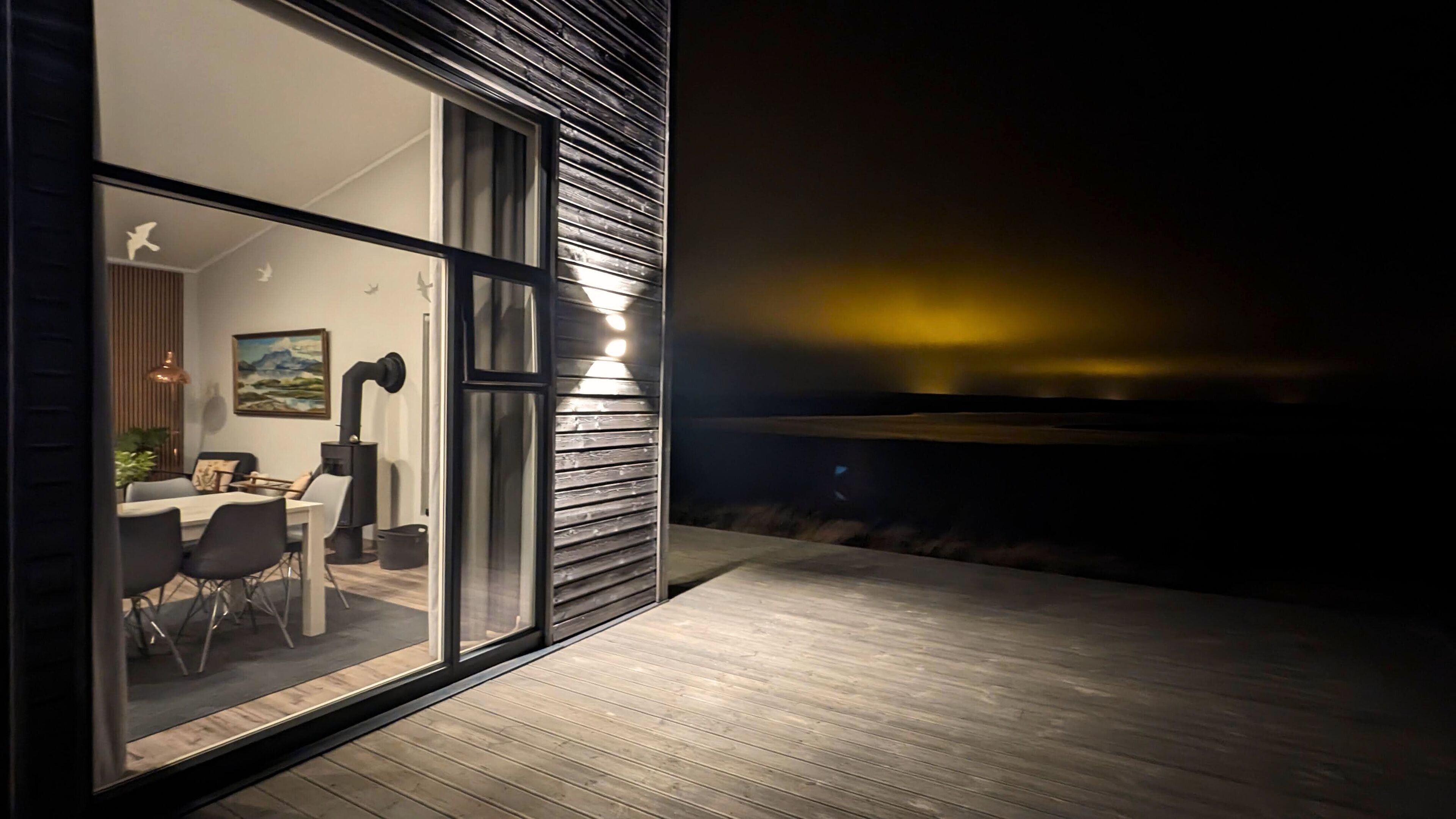 A modern outdoor dining area with a glass wall, wooden deck, and a view of the night sky.