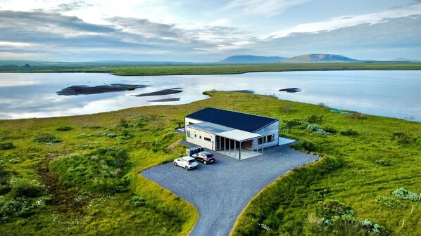 A modern house with a large garage and two cars parked outside, surrounded by a grassy landscape and a body of water. - Austurey Lakefront Villa (Bláskógabyggd)