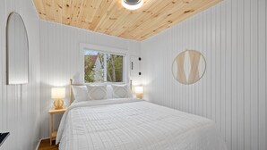 Ein Schlafzimmer mit Holzbalkendecke, weißen Wänden, einem großen Bett mit weißer Bettdecke, zwei Nachttischen mit Lampen und einem Fenster mit Blick auf Bäume.
