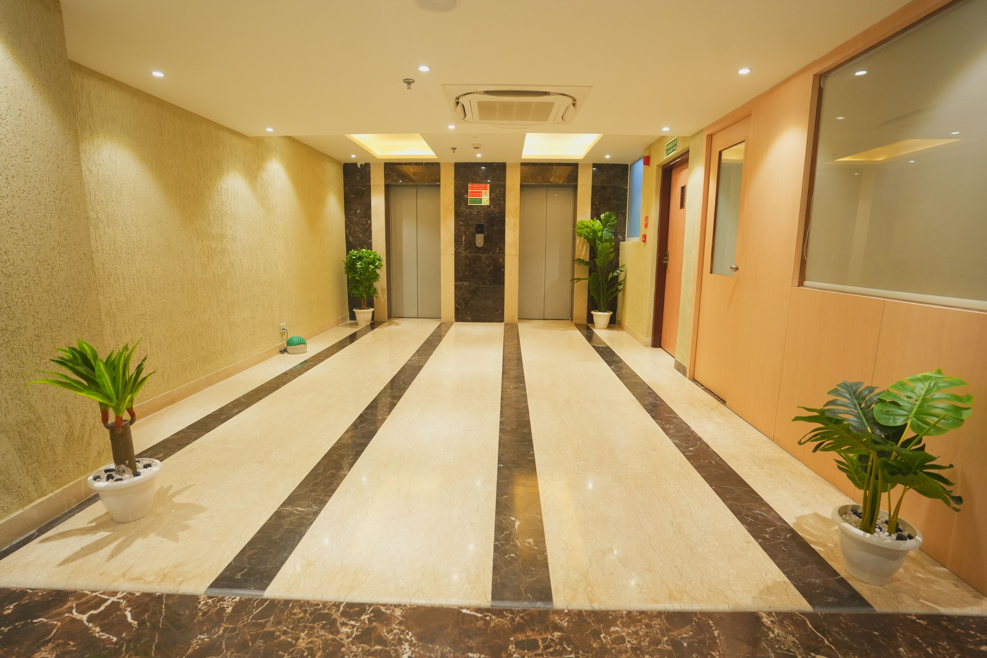 A hotel lobby with a marble floor, two elevators, potted plants, and a reception desk.