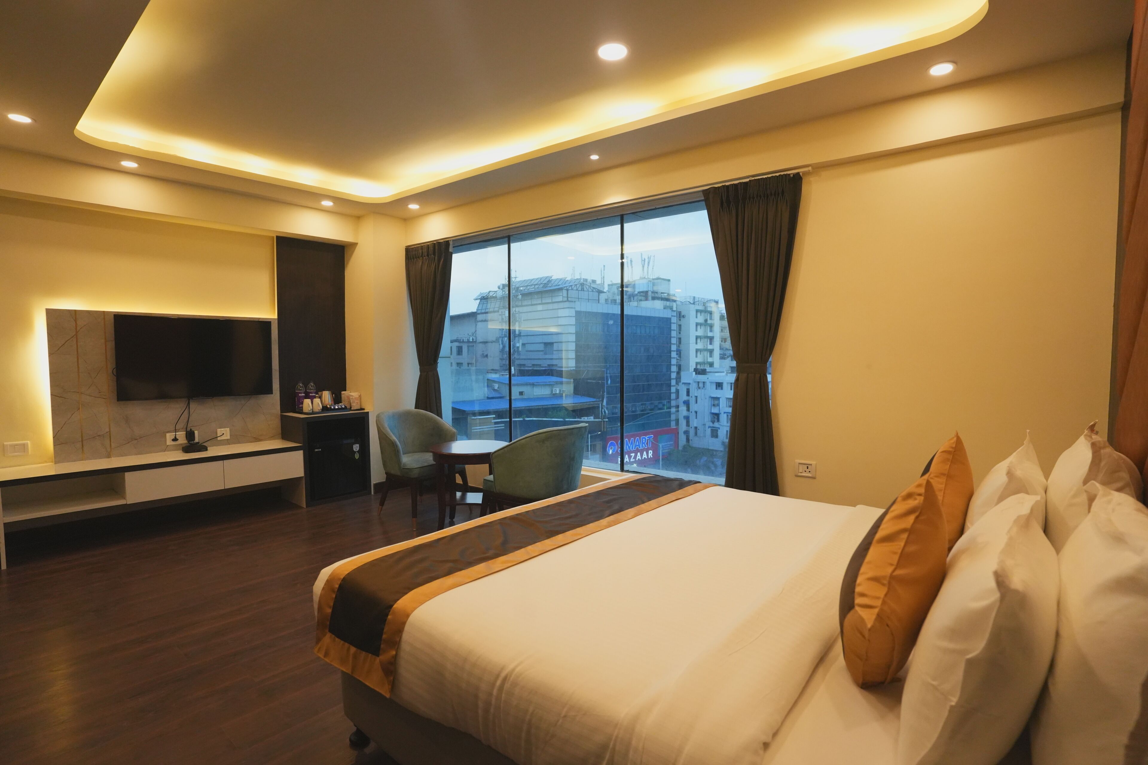 A modern hotel room with a large bed, a flat-screen TV mounted on the wall, a desk with a chair, and a large window offering a city view.