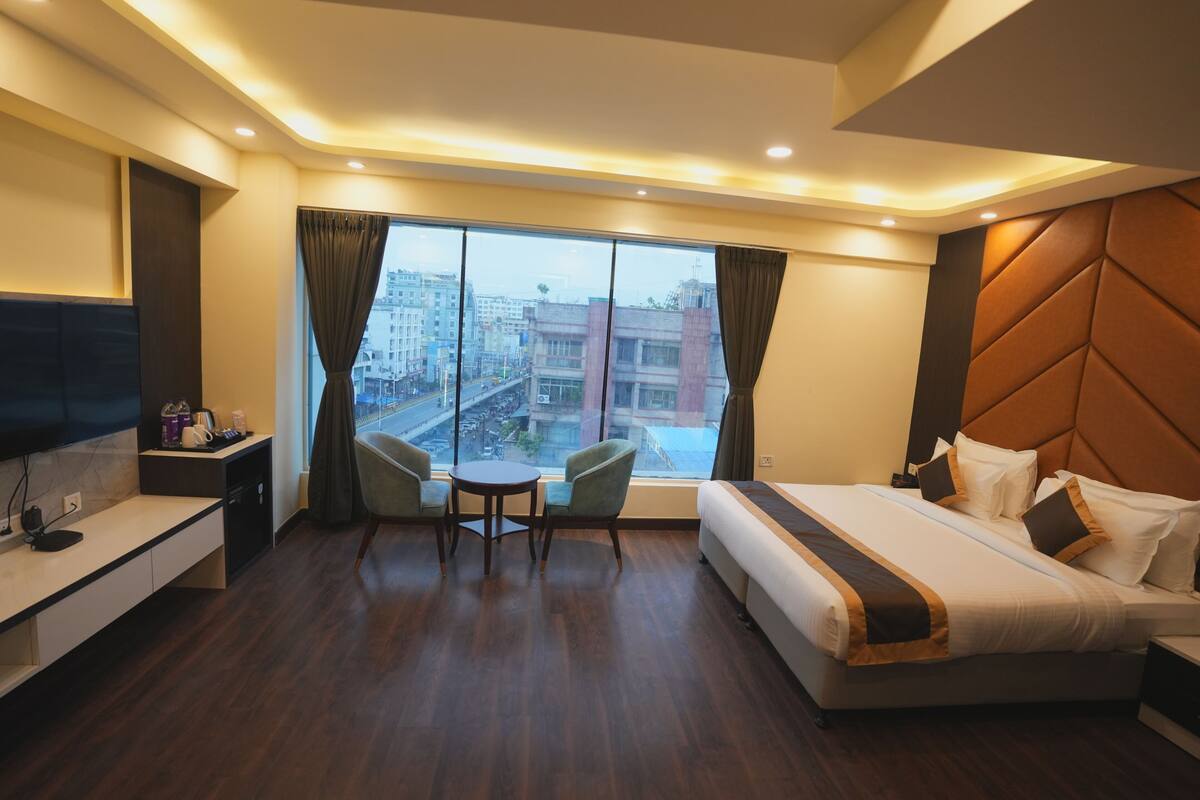 Habitación de hotel con una cama grande, un escritorio con televisor de pantalla plana, una mesita con dos sillas y un ventanal con vistas a la ciudad.