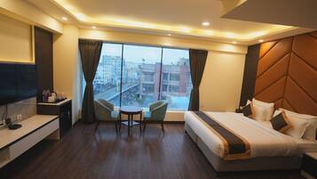 A hotel room with a large bed, a desk with a flat-screen TV, a small table with two chairs, and a large window with a city view.
