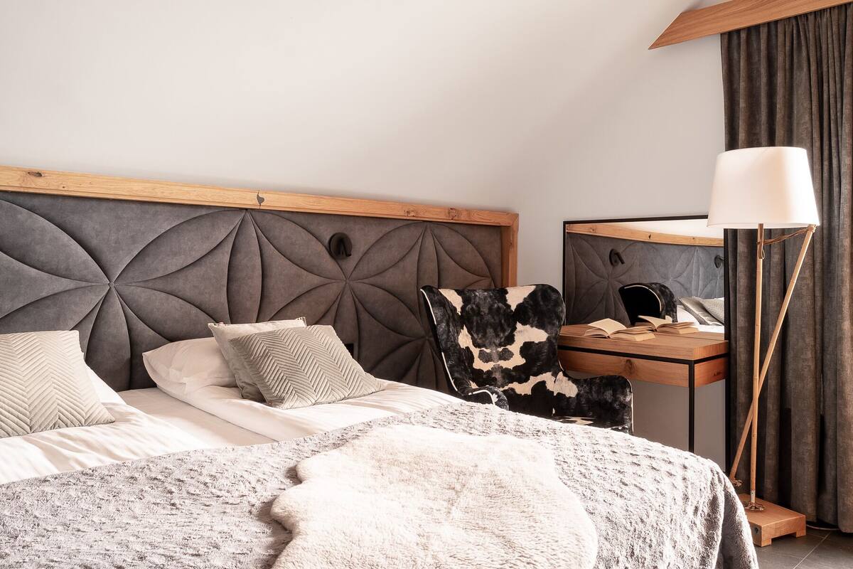 Une chambre moderne avec une tête de lit capitonnée, un grand lit avec de nombreux oreillers, une table de chevet avec une lampe et une chaise agrémentée d’un coussin à motif de peau de vache.
