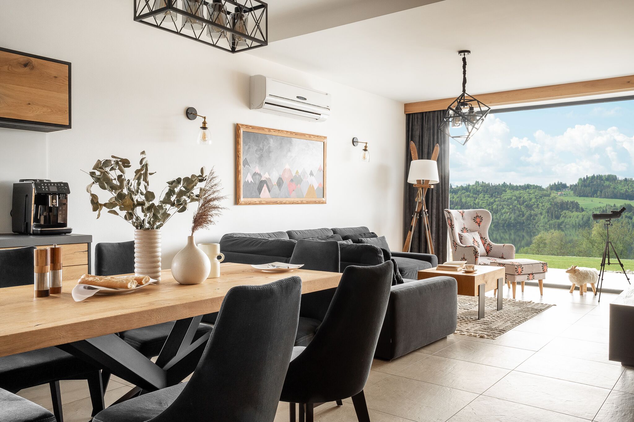 Een moderne woonkamer met een houten eettafel, zwarte banken en een groot raam met uitzicht op een groen landschap.