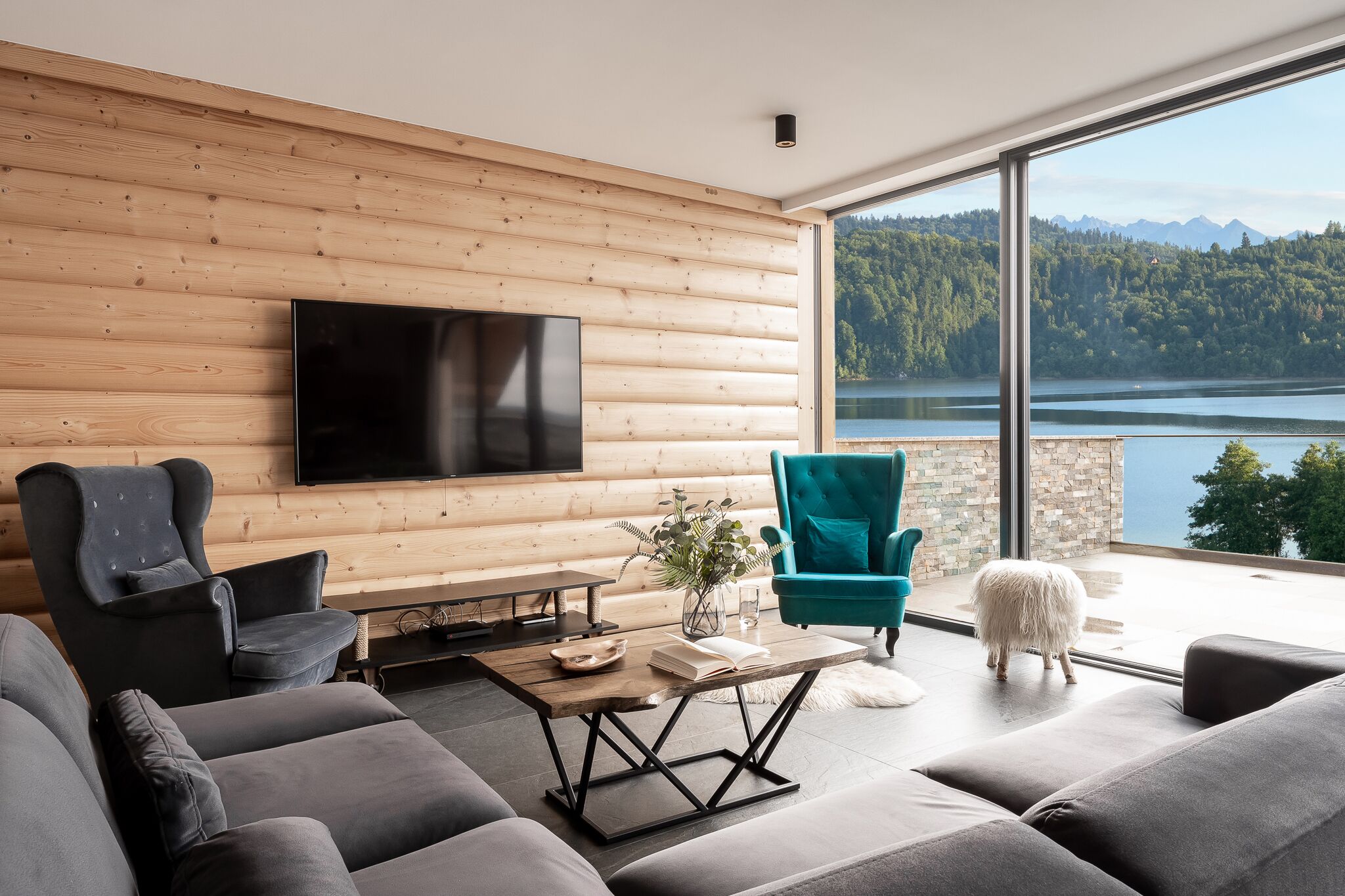Un salon moderne doté d’un grand téléviseur, de sièges confortables, d’un mur en bois et offrant une vue sur un lac et des montagnes.