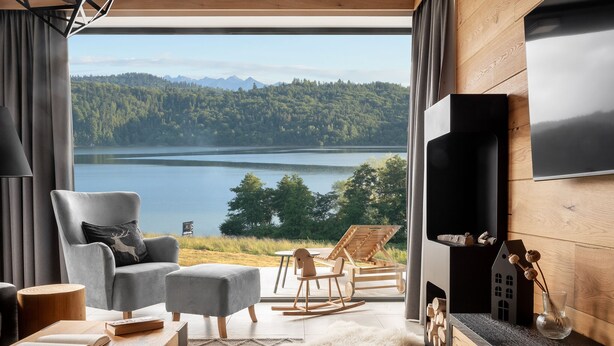 A modern living room with a large window overlooking a lake and forest, a comfortable seating area, and a flat-screen TV mounted on the wall. - Czorsztyn Prestige (Czorsztyn)