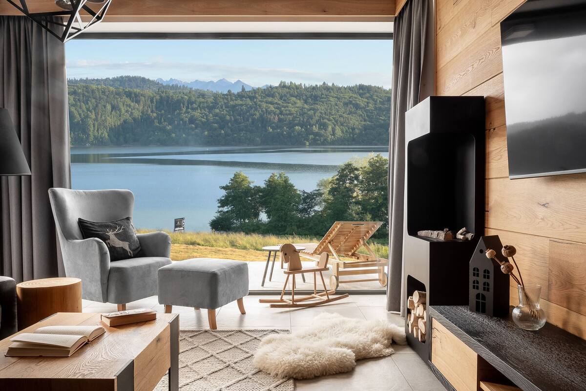 Un salon moderne doté d’une grande fenêtre donnant sur un lac et une forêt, d’un coin salon confortable et d’un téléviseur à écran plat fixé au mur.