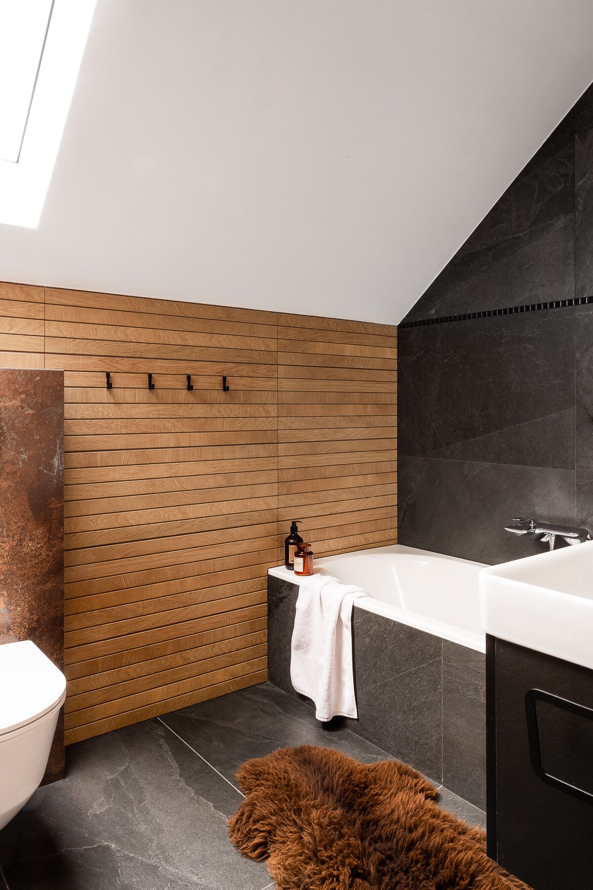 A modern bathroom with a wooden panel wall, a black marble wall, a white bathtub, and a fluffy brown rug.