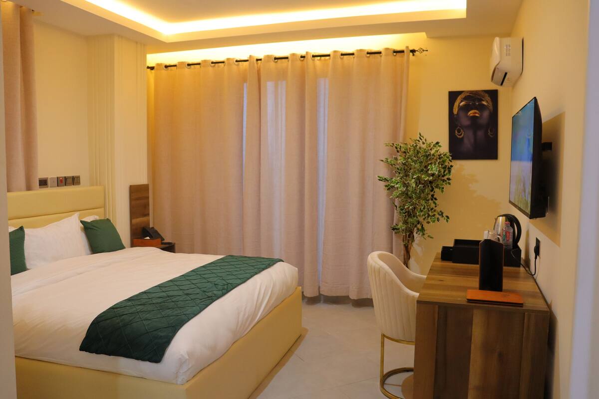 A hotel room with a bed, a desk, a chair, a plant, and a TV.