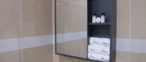A bathroom with a tiled wall, a mirror, and a shelf with toiletries and towels.