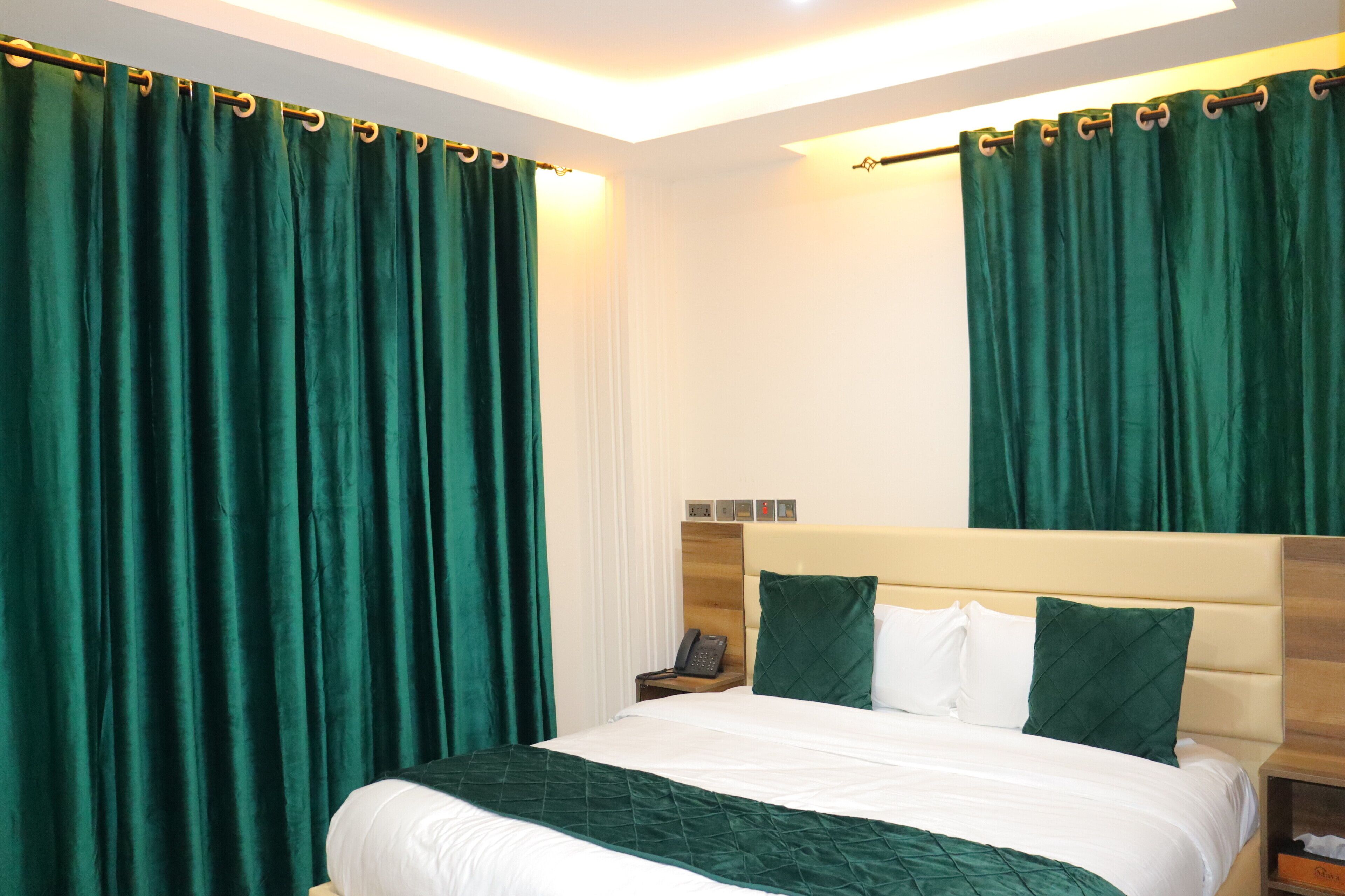 A hotel room with a large bed, green curtains, a wooden headboard, and a bedside table with a phone.