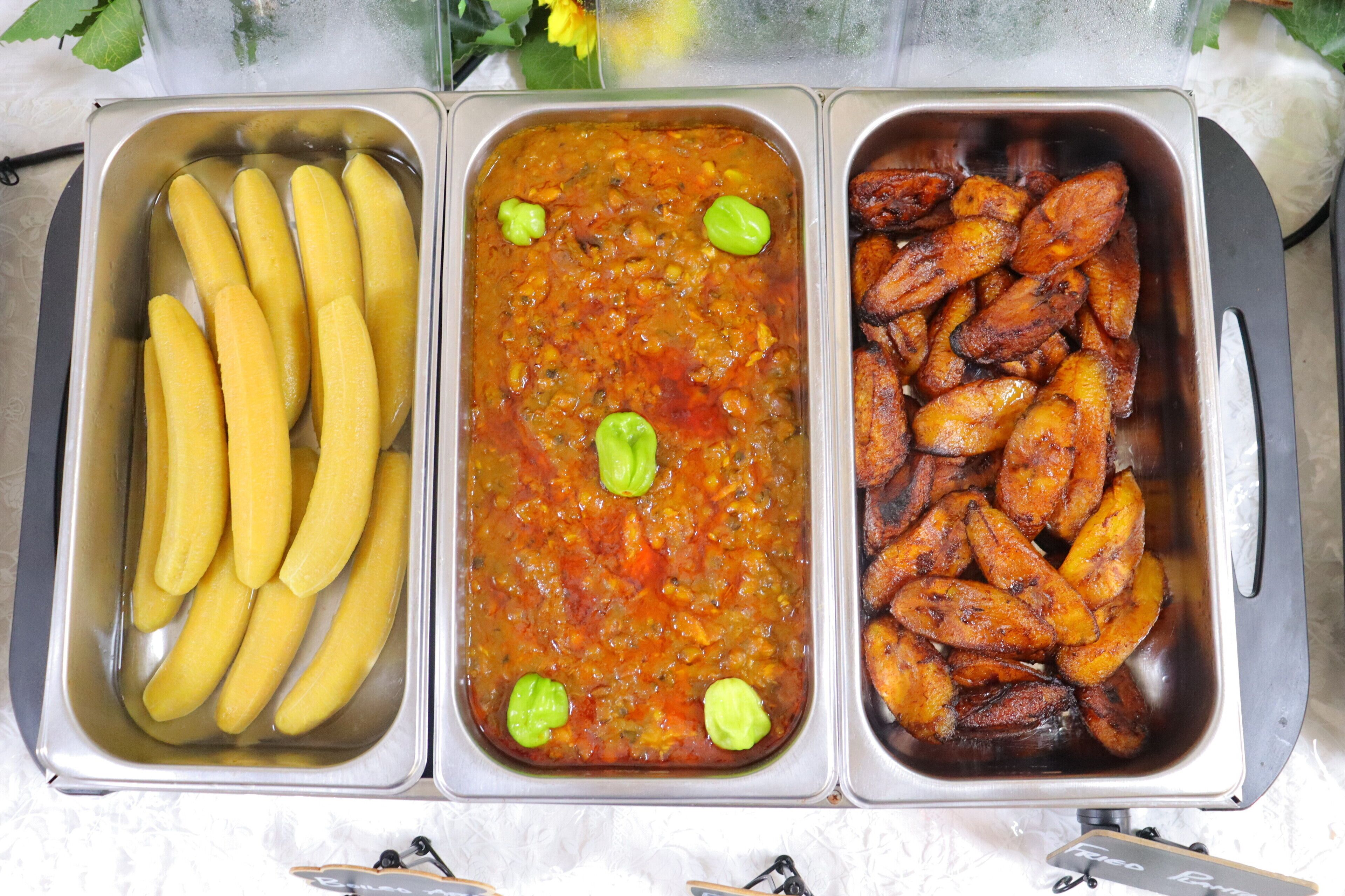 A buffet with three trays containing bananas, a vegetable dish, and roasted potatoes.
