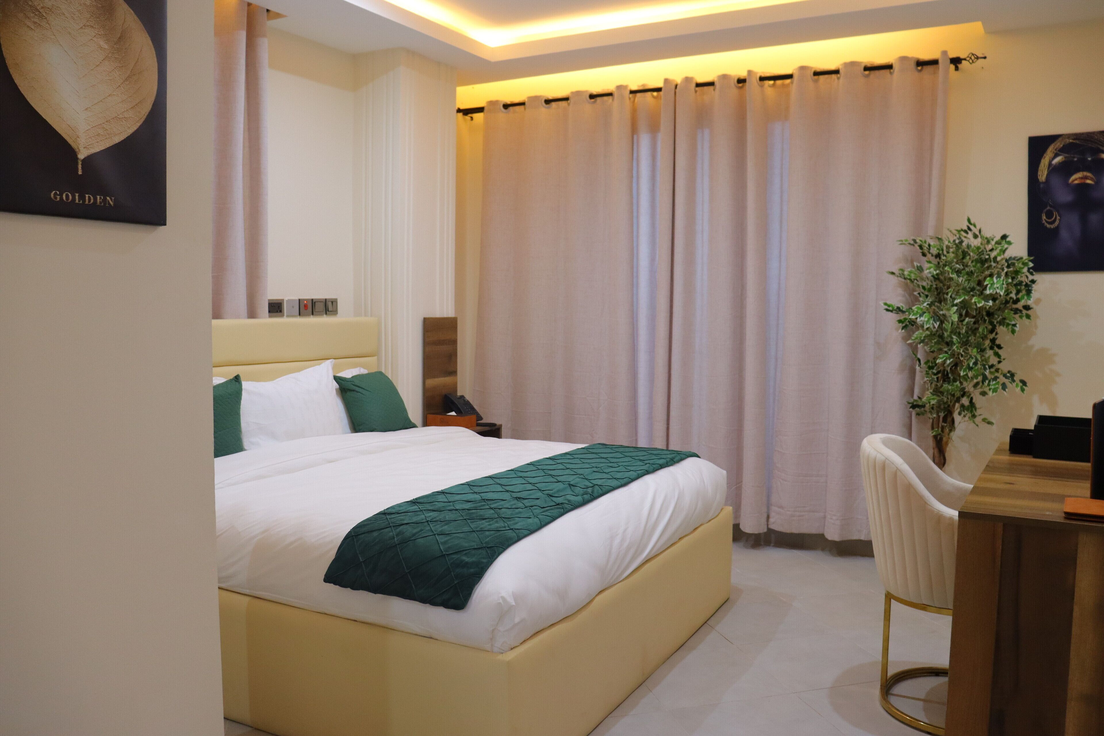 A hotel room with a bed, a desk, a chair, a plant, and a wall-mounted artwork.