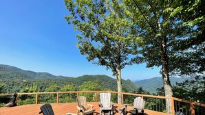Een houten terras met Adirondack-stoelen en een vuurkuil, met uitzicht op een bergachtig landschap.