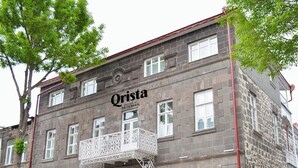 Un edificio histórico con el nombre 'Qrista' en la fachada, que presenta un balcón con ornamentación metálica, una alfombra roja en la entrada y paredes de piedra.