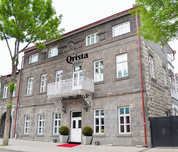 Un bâtiment historique dont la façade porte l’inscription « Qrista », avec un balcon orné de ferronnerie, un tapis rouge à l’entrée et des murs en pierre.