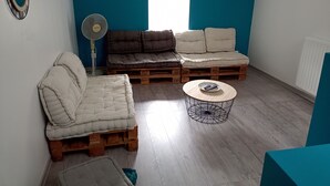 Een kamer met een blauwe accentmuur, een houten vloer, een witte bank, een bruine bank, een klein rond tafeltje en een staande ventilator.