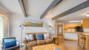 Een moderne woonkamer met een leren bank, een houten salontafel en een schilderij van een berglandschap. In de achtergrond is een keuken met houten kastjes en een koelkast zichtbaar.