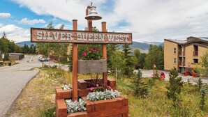 Een rustiek houten bord met de tekst 'Silver Creek West' en een bel bovenop, omgeven door bloembakken en een bergachtig landschap.
