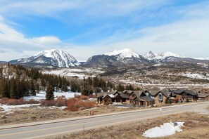 A row of log cabin-style buildings with a road in the foreground and snow-covered mountains in the background.