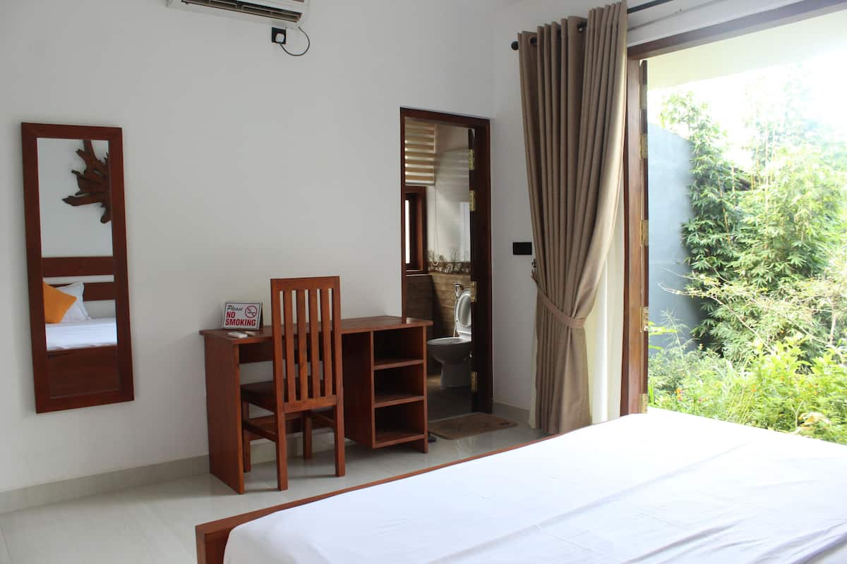 A bedroom with a bed, a desk, and a chair. There is a sign on the desk that says "Please No Smoking." The room has a window with curtains, and a view of greenery outside.
