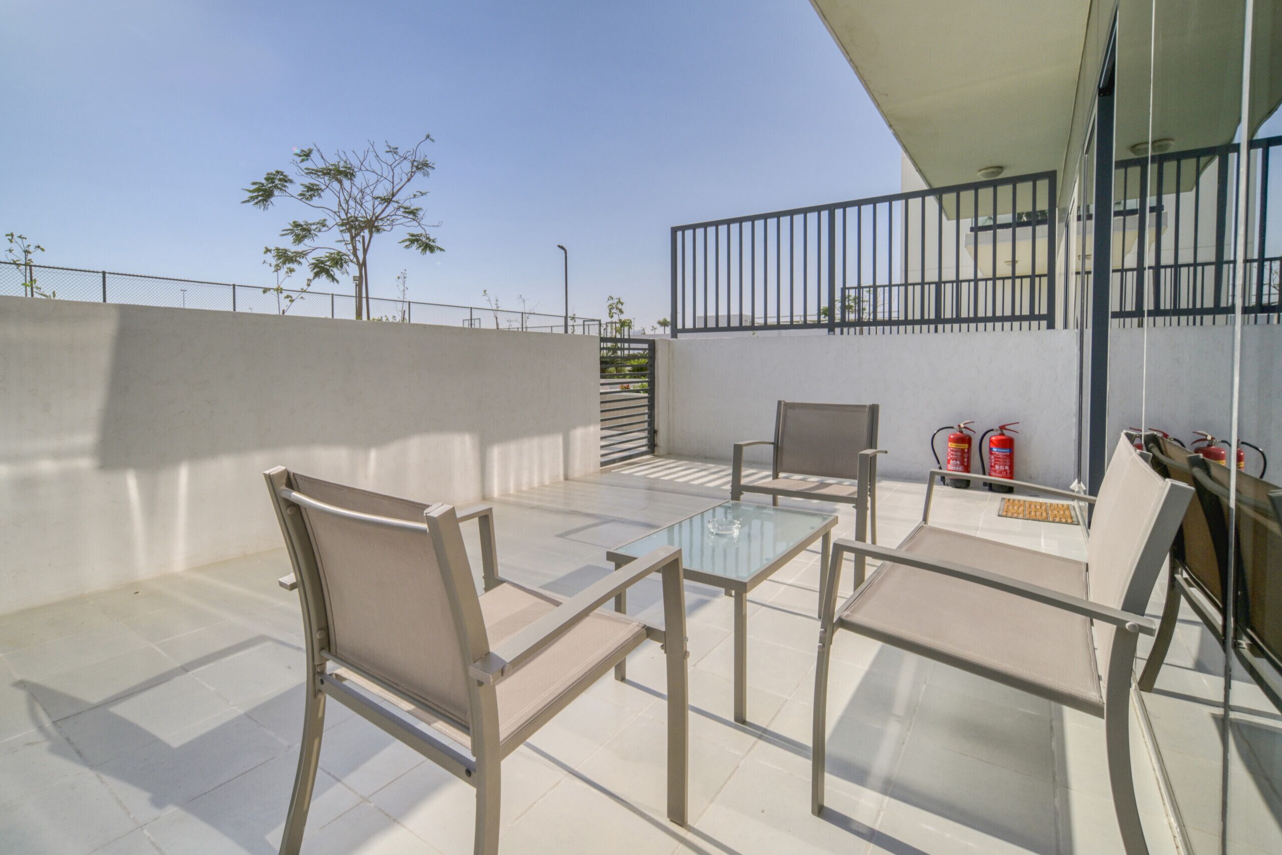 An outdoor patio with a glass table and chairs, a fire extinguisher, and a railing.