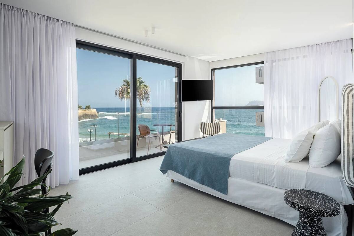 Quarto de hotel moderno com uma cama grande, televisão e varanda vista para o oceano.