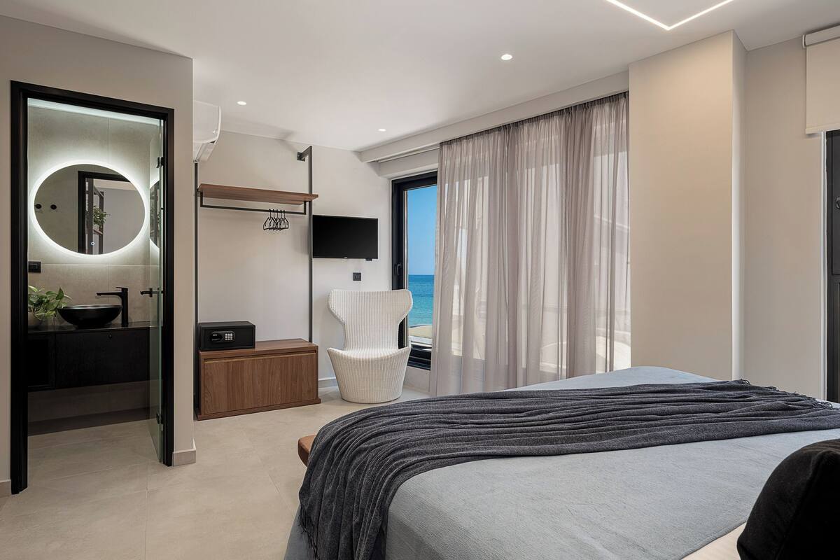 Quarto moderno com uma cama grande, banheiro com espelho redondo, televisão e vista para o mar.