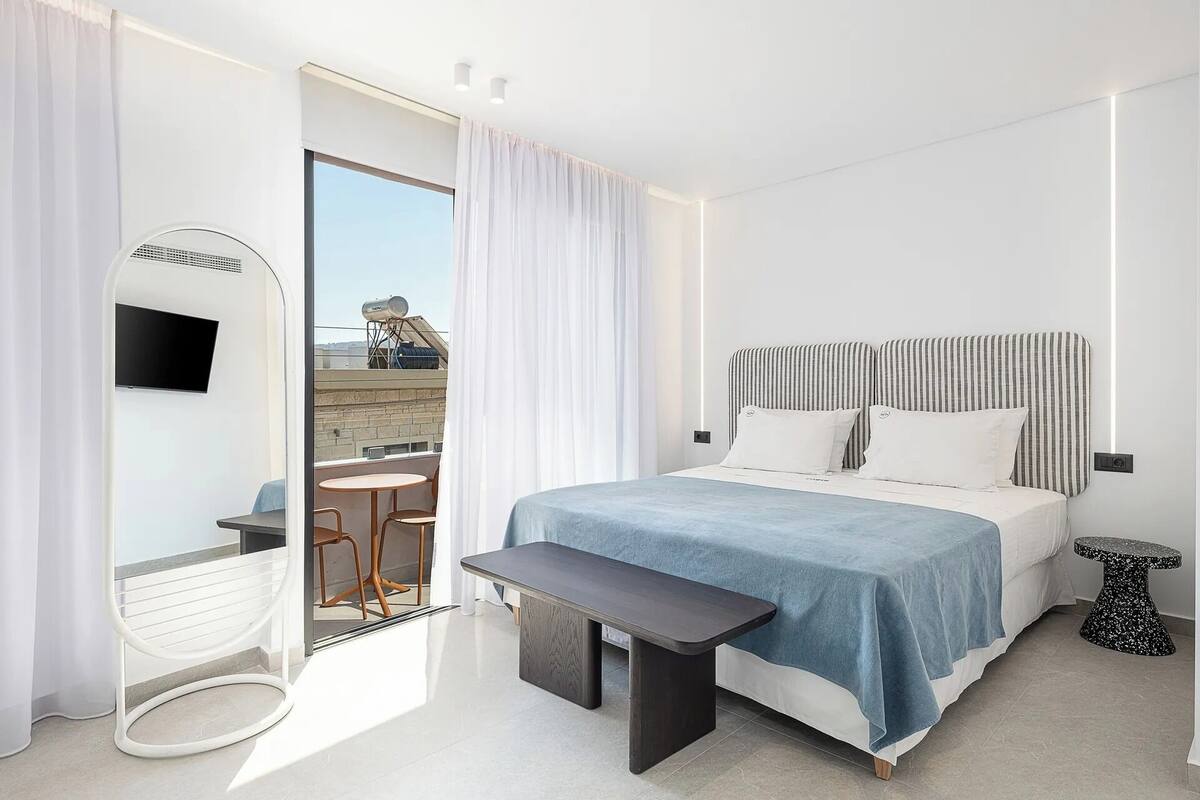 Quarto de hotel moderno com cama, uma mesa pequena, um banco, televisão e vista para o exterior.