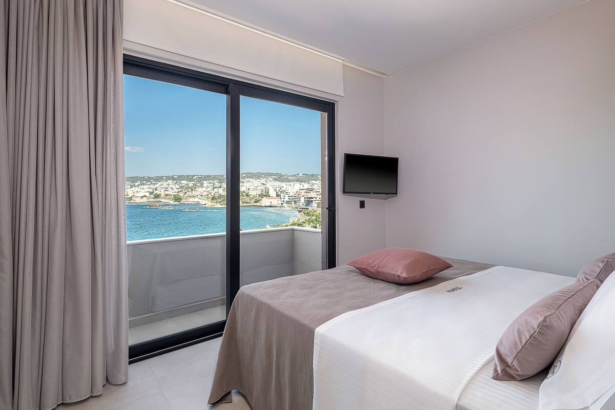 Quarto de hotel com uma grande janela, uma cama, uma televisão fixada na parede e vista para uma cidade costeira.