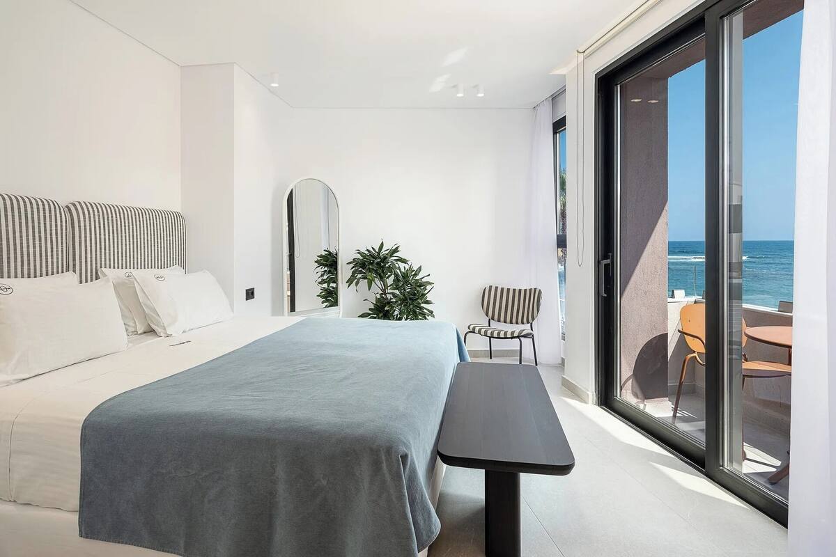 Quarto moderno com uma cama grande, uma mesinha, uma cadeira, um espelho e vista para o mar através de uma porta de correr de vidro.