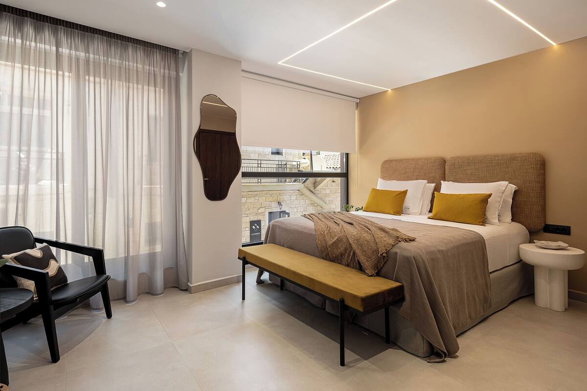 Quarto moderno com cama grande, um banco, mesa de cabeceira e vista para o exterior.