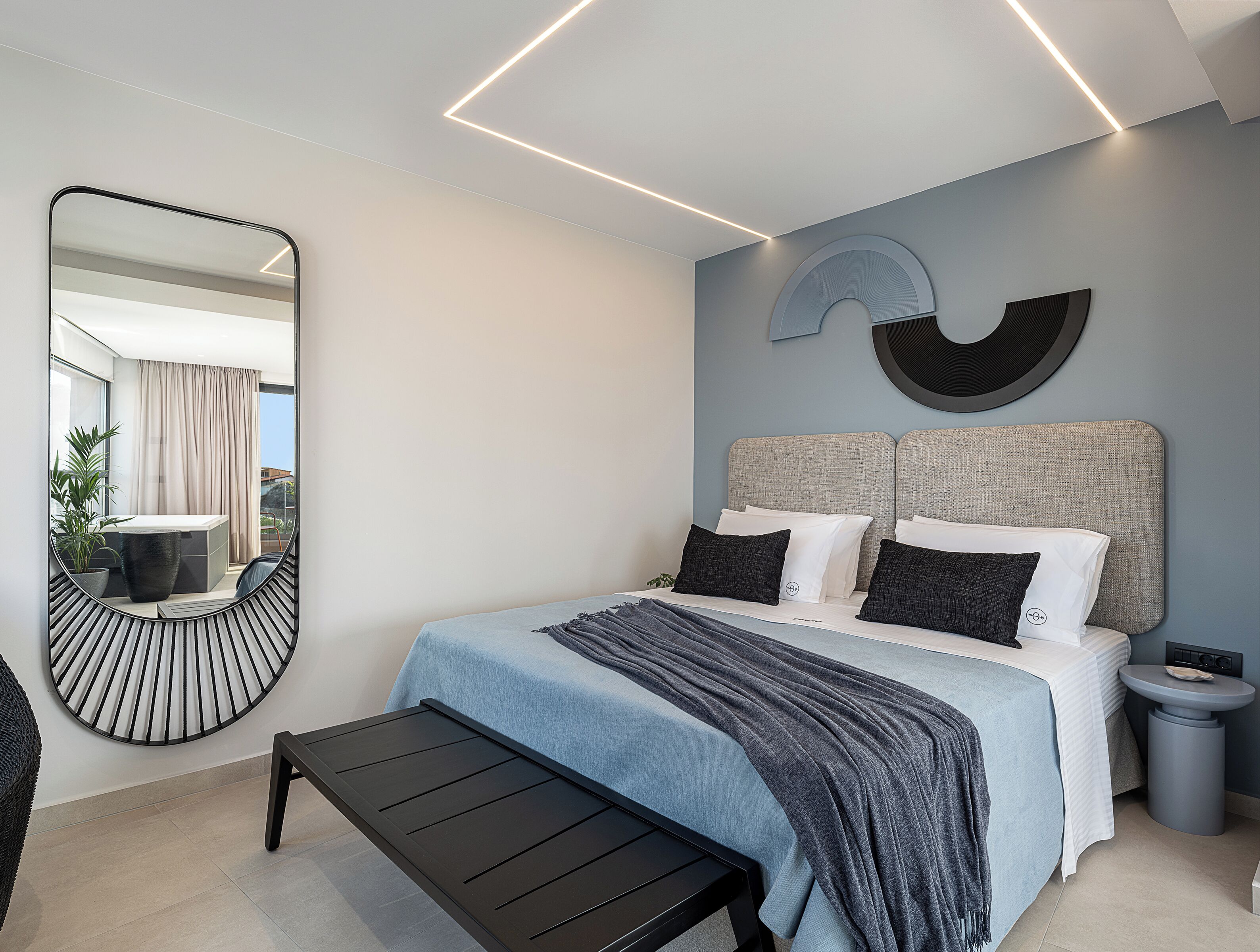 Un dormitorio moderno con una cama, mesitas de noche, un banco, un espejo y una obra de arte en la pared.