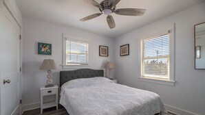 Un dormitorio con una cama, dos mesitas de noche, un ventilador de techo y dos ventanas con persianas.