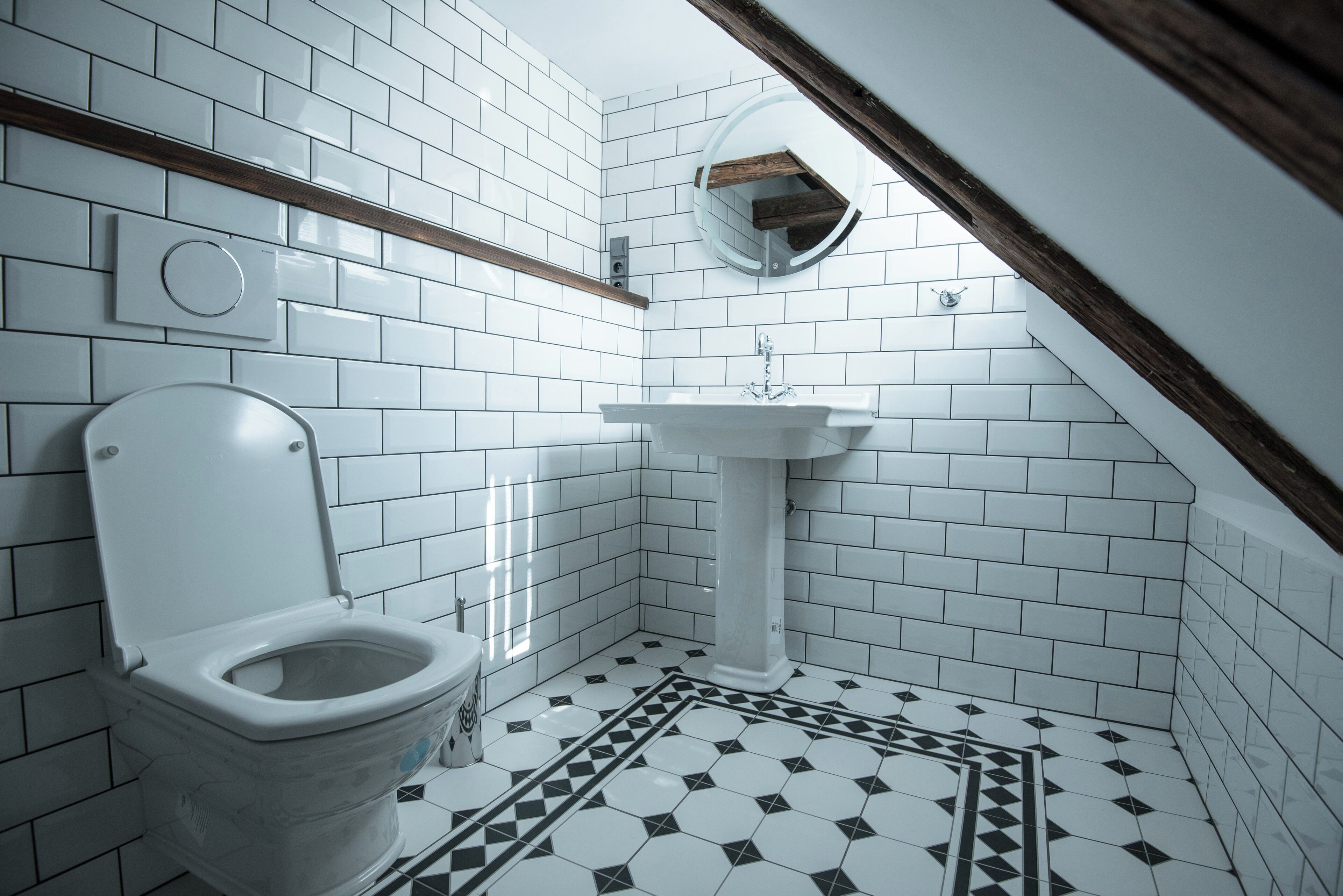 Une salle de bain avec des murs recouverts de carreaux blancs, des toilettes blanches, un lavabo blanc et un sol à motifs.