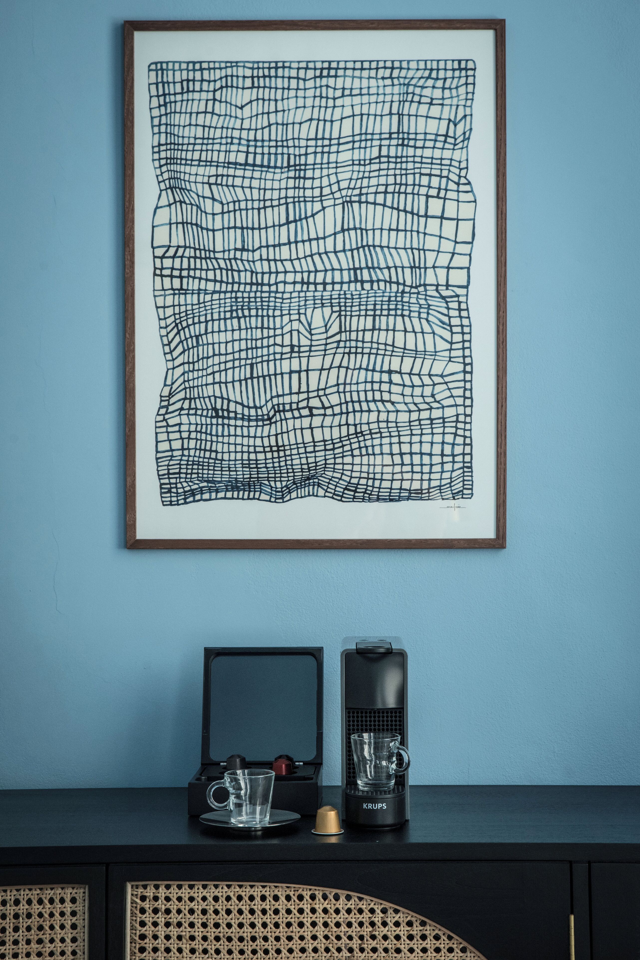 A framed abstract artwork on a wall, a coffee maker, and a glass on a dark surface.