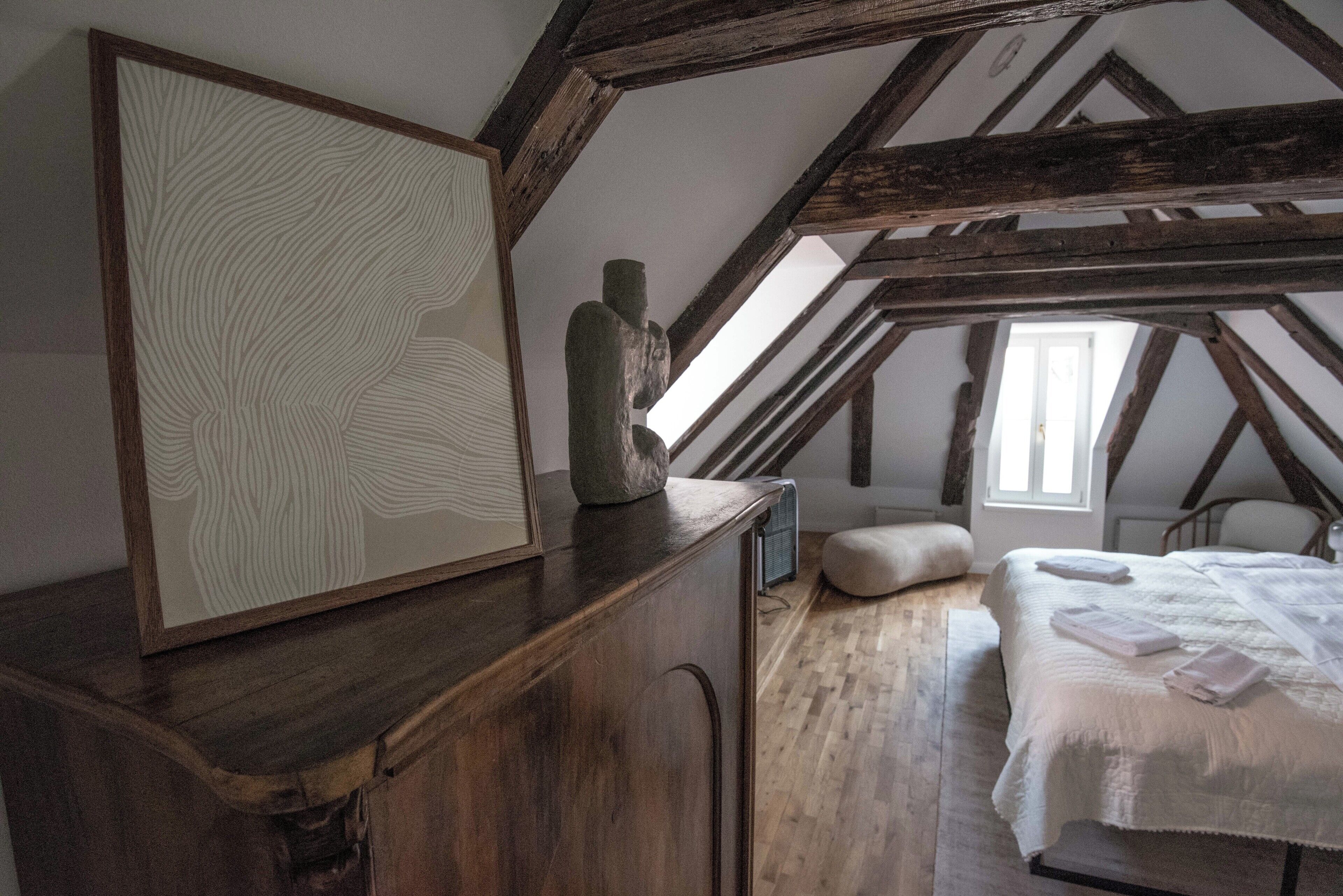 Un dormitorio con techo inclinado, vigas de madera, una cama con ropa blanca y una obra de arte enmarcada sobre un aparador de madera.