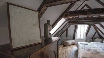 Un dormitorio con techo inclinado, vigas de madera, una cama con ropa blanca y una obra de arte enmarcada sobre un aparador de madera.