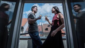 Um casal brindando com taças de vinho em um quarto com vista para prédios e céu azul.
